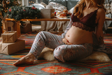 Beautiful Pregnant Woman In Comfy Clothes sitting on a flor Near The Christmas Tree.