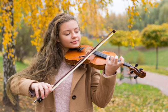 Girl playing the violin in the autumn park on a background of yellow foliage. Young violinist playing in the park