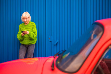 Colors and alternative happy lifestyle concept image with blonde attractive woman thirty years old using mobile phone internet technology outside the red car and with blue wall on the background