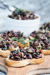 Homemade mixed Olive Tapenade made with garlic, capers, olive oil, Kalamata, black and green olives spread over toasted bread. Selective focus on canape in foreground with blurred background.