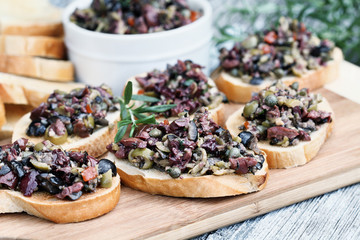 Homemade mixed Olive Tapenade made with garlic, capers, olive oil, Kalamata, black and green olives spread over toasted bread. Selective focus on center canape with blurred background.