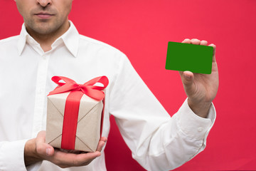 Attractive businessman in white shirt showing business card to the camera, holding a box with a gift with a red ribbon, bright red background, with copy space, for advertisement, front view, close up