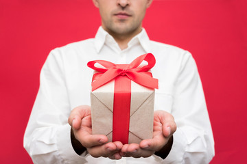 Attractive man in white shirt holding a box with a gift with a red ribbon, on camera, bright red background, front view, close up