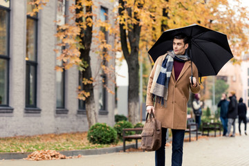 handsome stylish man in coat and scarf with umbrella walking by autumnal street