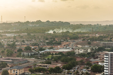 Obraz premium High view point cityscape of a neighborhood in de hills of Accra, Ghana. Apartment buildings, slums, construction sites, wastelands and streets . Smoke from burning waste with a green and hazy horizo