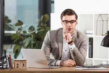 Naklejka premium handsome confident businessman sitting at workplace and looking at camera in modern office