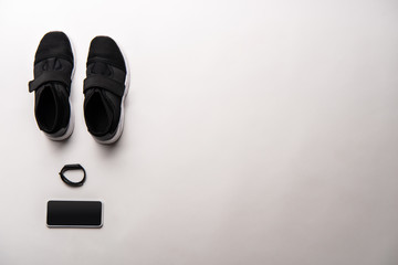view from above of arranged black sport shoes, fitness tracker and smartphone with blank screen isolated on white