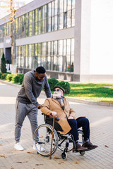 senior disabled man in wheelchair with walking stick and african american cuidador talking on street