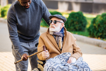 young african american man giving walking stick to senior disabled man in wheelchair on street