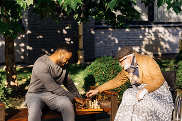 senior disabled man in wheelchair and african american nurse playing chess together on street