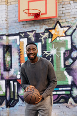 happy african american man with basketball ball looking at camera on street in front of colorful graffiti