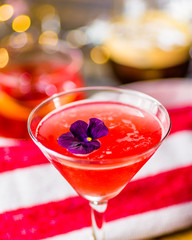 A red colored cocktail with a purple flower, with other cocktails in the background  