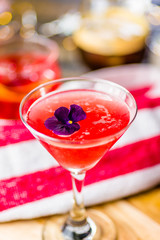 A red colored cocktail with a purple flower, with other cocktails in the background  
