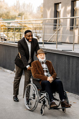happy senior disabled man in wheelchair and african american man riding by street