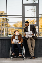 senior disabled man in wheelchair and african american man reading newspapers together on street