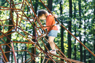 Naklejka na ściany i meble Active little child playing on climbing net the playground. A girl play and climb outdoors on sunny summer day. 