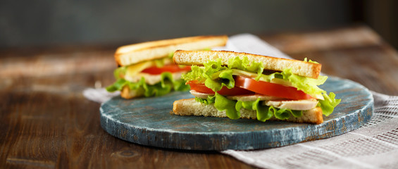 Appetizing sandwich with chicken, tomatoes, lettuce, cheese on a wooden plate on a dark background
