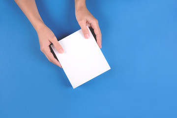 Feminine hands holding white gift box on blue paper background. Top view. Copy space. Holiday celebration or skin care concept. Toned