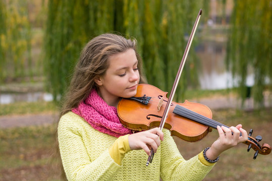 Girl playing the violin eyes closed in the autumn park at a lake and willow foliage background.