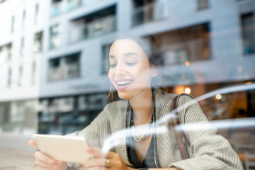 View through the window on the stylish woman sitting with phone indoors with modern building on the reflection