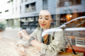 Fototapeta premium View through the window on the stylish woman sitting with phone indoors with modern building on the reflection