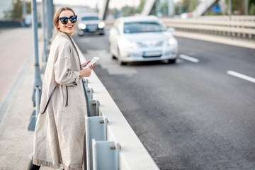 Obraz premium Lifestyle portrait of a young stylish woman with bag and phone outdoors on the modern bridge