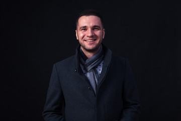 Obraz premium Młody brunet z brązowymi oczami w studiu na czarnym tle i białym dymie, ubrany w ciemnoszary płaszcz, szalik w kratkę. Koncepcja zimnej pogody, para z ust, początek zimy
