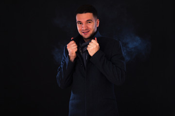 Young male brunette with brown eyes in the studio on a black background and white smoke, dressed in a dark gray coat, checkered scarf. Cold weather concept, steam from mouth, beginning of winter