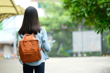 Back of student girl holding books and carry school bag while walking in school campus background,...