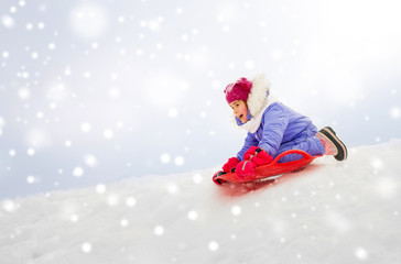 childhood, sledging and season concept - happy little girl sliding down on snow saucer sled outdoors in winter
