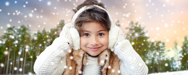 winter, people and christmas concept - happy little girl wearing earmuffs over snowy forest...