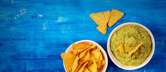 Fresh guacamole sauce in bowl with Mexican corn chips over bright blue background, top view with...