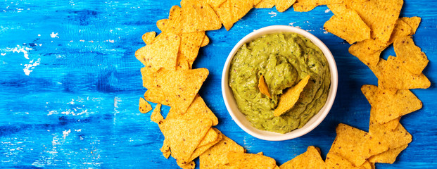 Fresh guacamole sauce in bowl with Mexican corn chips over blue background, top view.