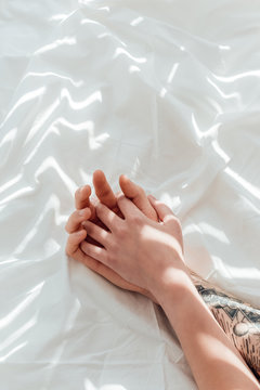 partial view of loving couple holding hands while lying on white bed sheet