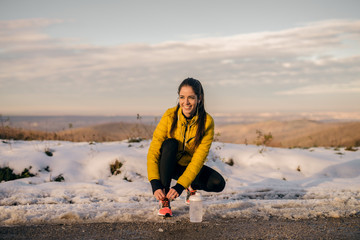 Happy sporty woman crouching and tying shoelace on the road in. In front of her bottle with water,...