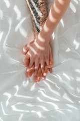partial view of loving couple holding hands while lying on white bed sheet