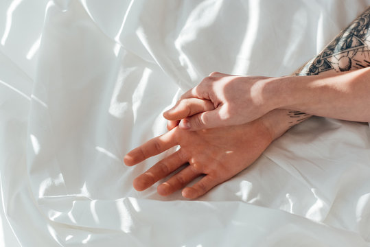 partial view of loving couple holding hands while lying on white bed sheet