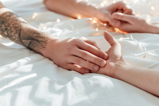 partial view of couple holding hands while lying in bed with garland