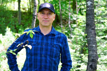 A caucasian happy man is looking into the camera for a portrait while hiking in the forest wearing a blue check shirt and a black hat.