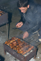 Mature man is cooking barbecued meat on mangal with skewers.