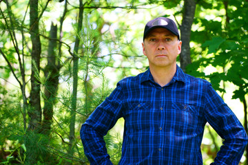 Naklejka na ściany i meble A caucasian serious man is looking into the camera for a portrait while hiking in the forest wearing a blue check shirt and a black hat.