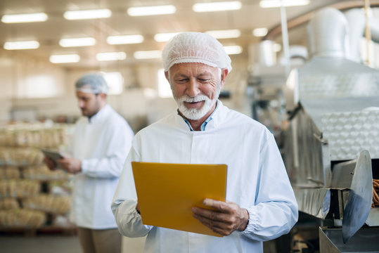 Senior worker checking documentation while standing in food factory. Protective uniform on.