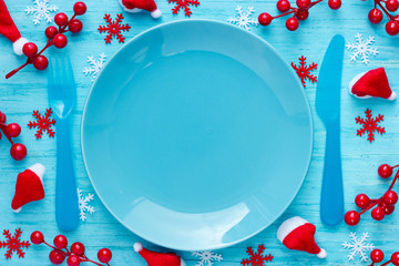 Christmas table place setting with empty blue plate and cutlery on blue background with festive decorations santa hat, snowflakes, decorative red berry. Christmas Xmas New Year holiday background