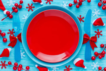 Christmas table place setting with empty red plate and cutlery on blue background with festive decorations santa hat, snowflakes, decorative red berry. Christmas Xmas New Year holiday background