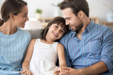 Attractive positive family with little daughter sitting together on sofa at home. Adorable girl smiling looking at camera loving mother and father holds her hands. Happy multi-ethnic family concept