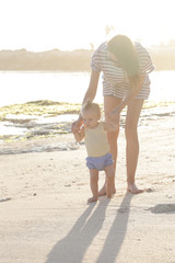 Mother helping her baby with the first steps, teaching baby to walk concept, outdoor candid photo on the beach, spending a day in the nature, healthy family lifestyle