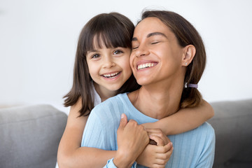 Close up portrait mom and daughter sitting on sofa at home. mum with closed eyes smiling her child...