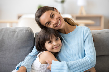 Head shot portrait smiling mother embrace little daughter sitting together on sofa posing looking at camera at home happy motherhood love and tenderness warm relationships between kid and mom.