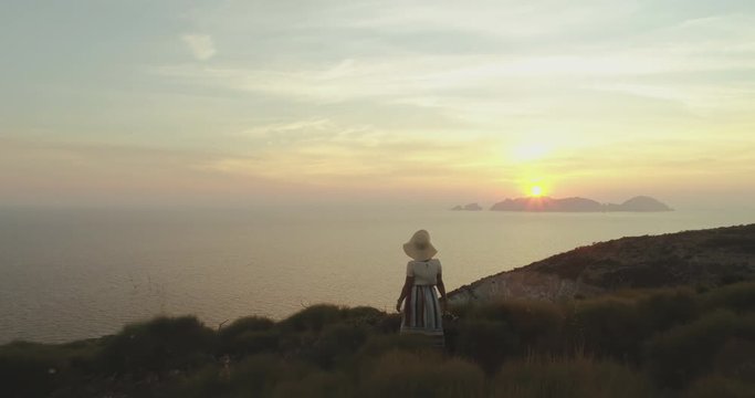 Beautiful young woman with hat in fashion colorful dress with skirt and flowers looking at the sunset on Ponza Island mountain Italy. Aerial Drone shot.