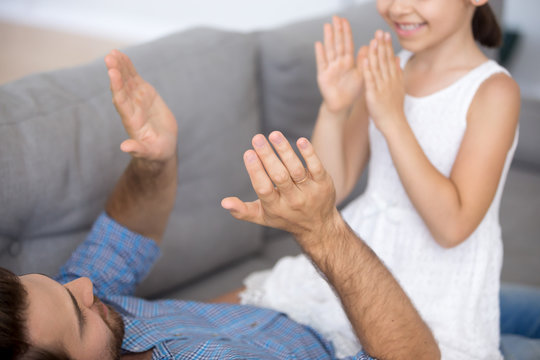 Little daughter and father playing pat-a-cake game clapping hands. Father lying on couch having fun together with child at home, close up focus on dad hands. Family weekend leisure activities concept